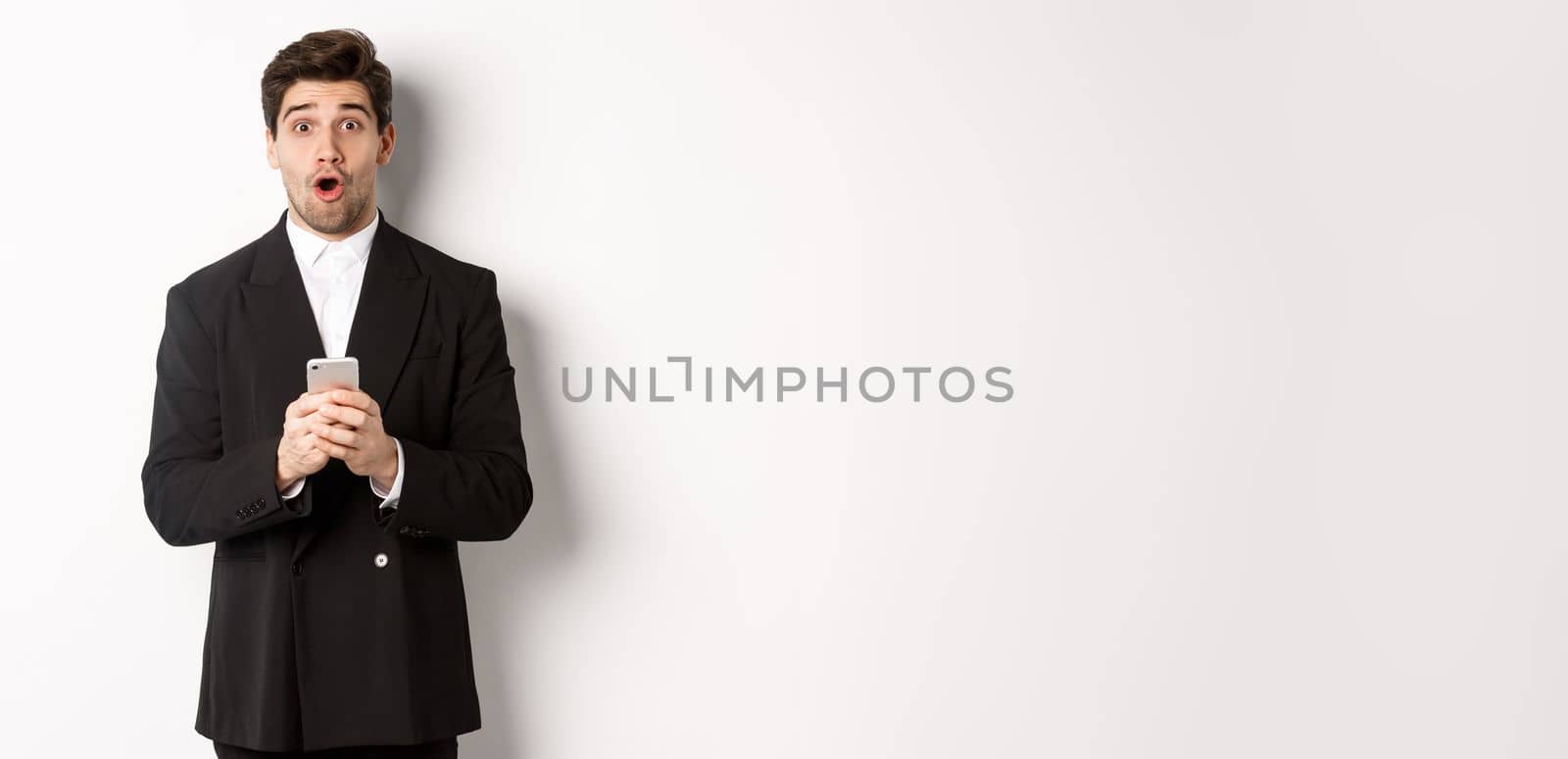 Amazed handsome guy in black suit reacting to cool promo offer, holding mobile phone, standing against white background.