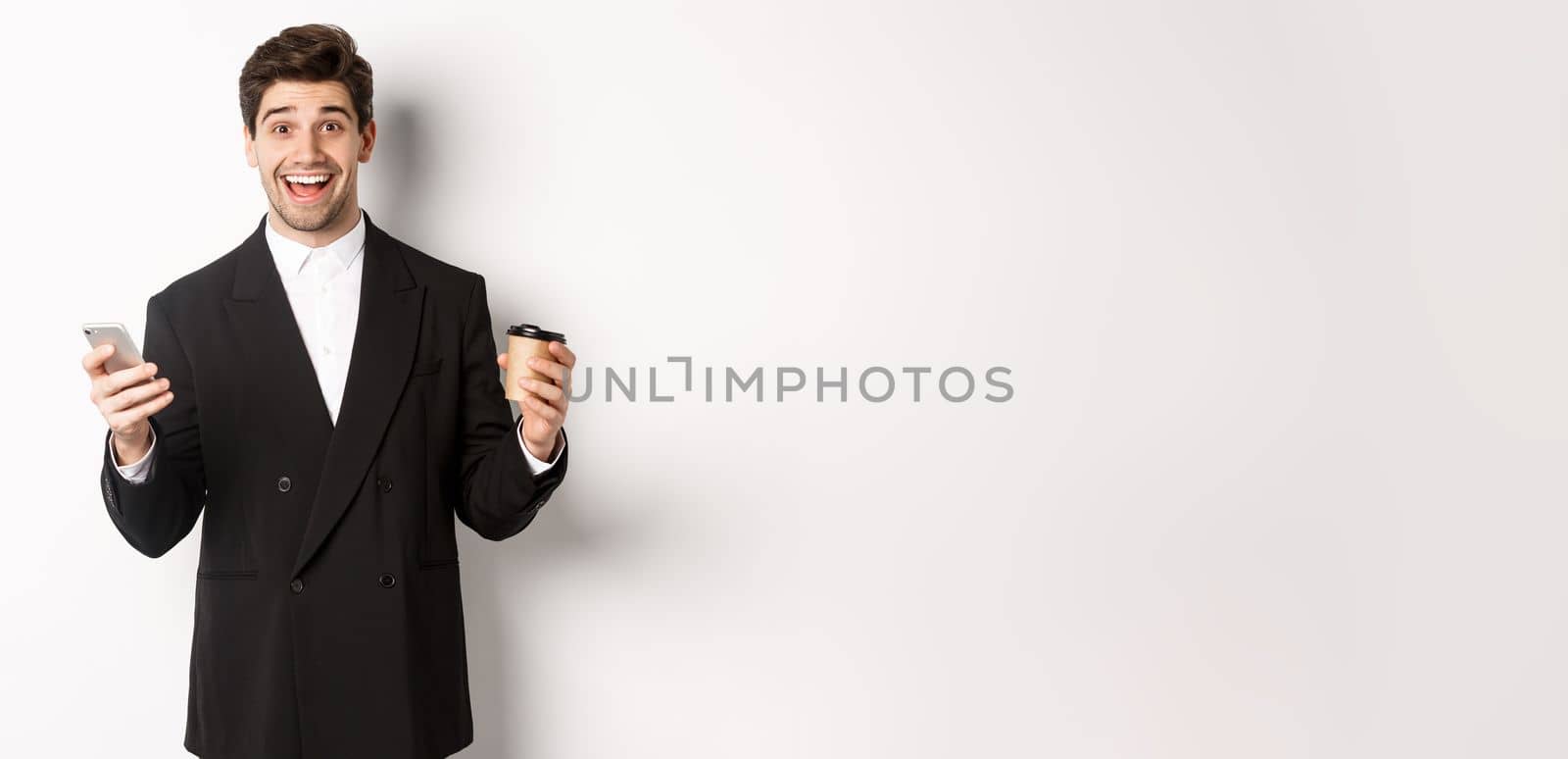 Portrait of happy good-looking man in suit, holding cup of coffee and smartphone, achieve app goal, standing over white background.