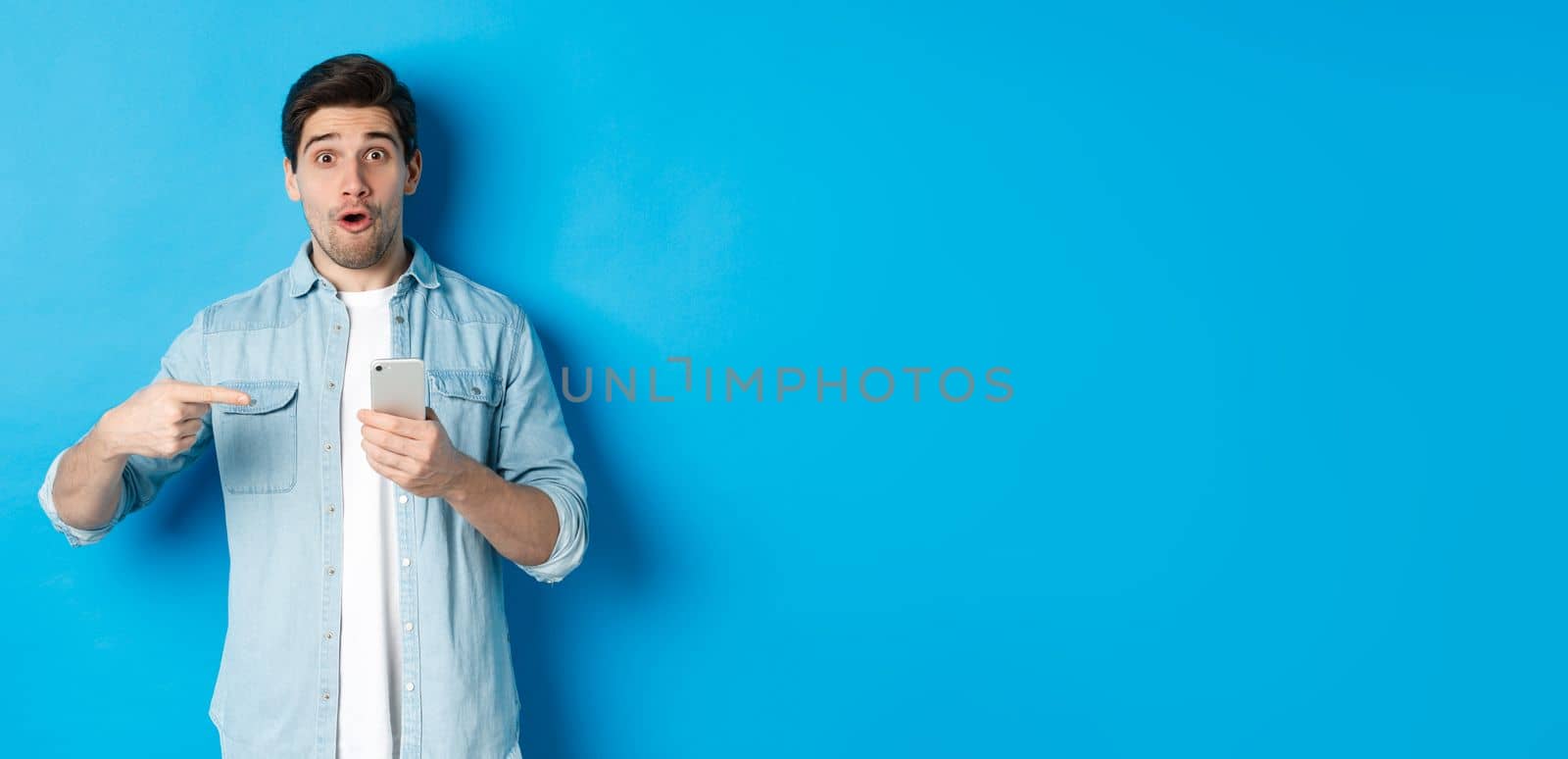 Concept of online shopping, applications and technology. Impressed man pointing finger at mobile phone and looking amazed, recommending app, standing over blue background.
