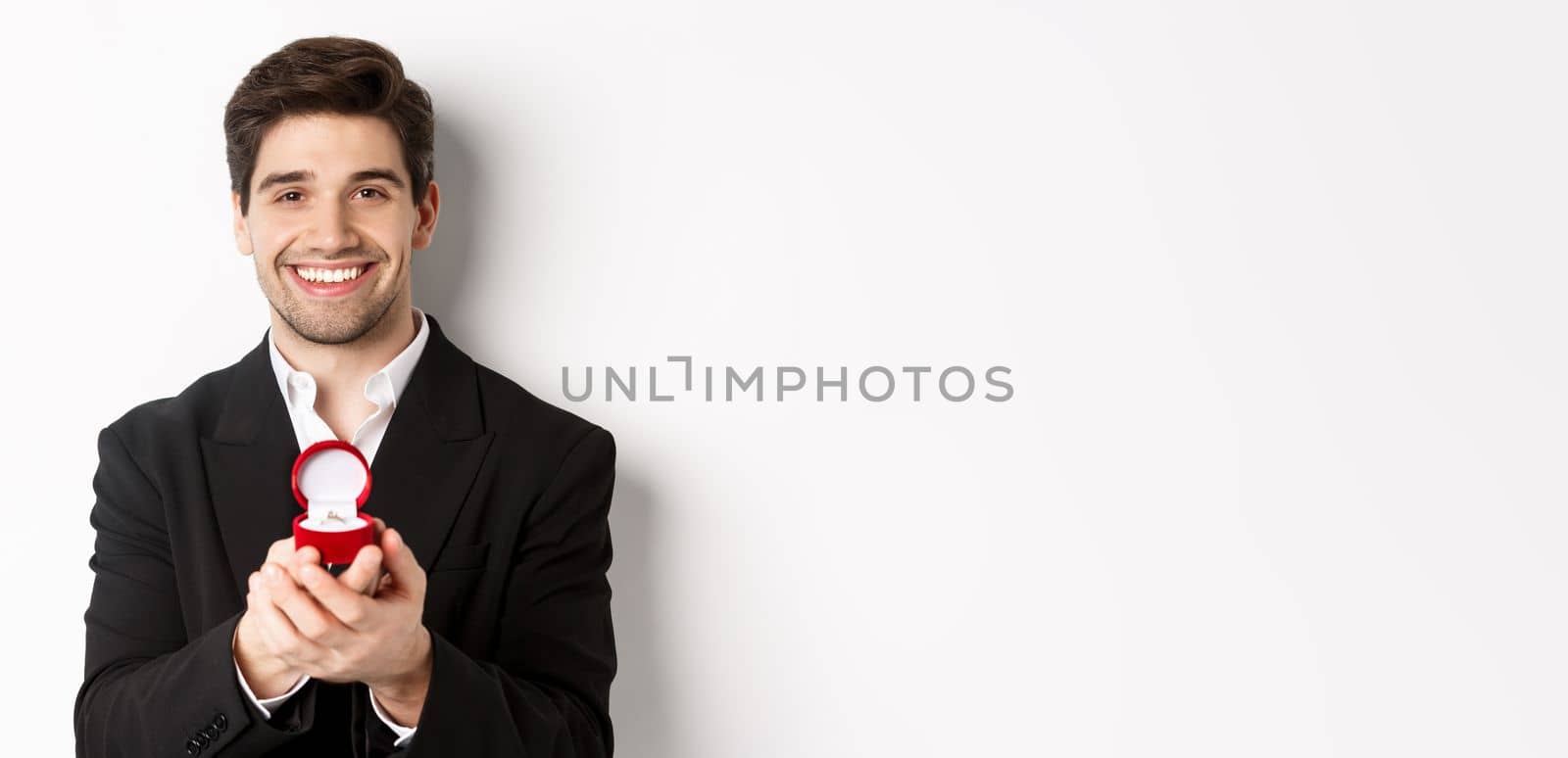 Image of handsome man looking romantic, open small box with engagement ring, making a proposal and smiling, standing against white background.