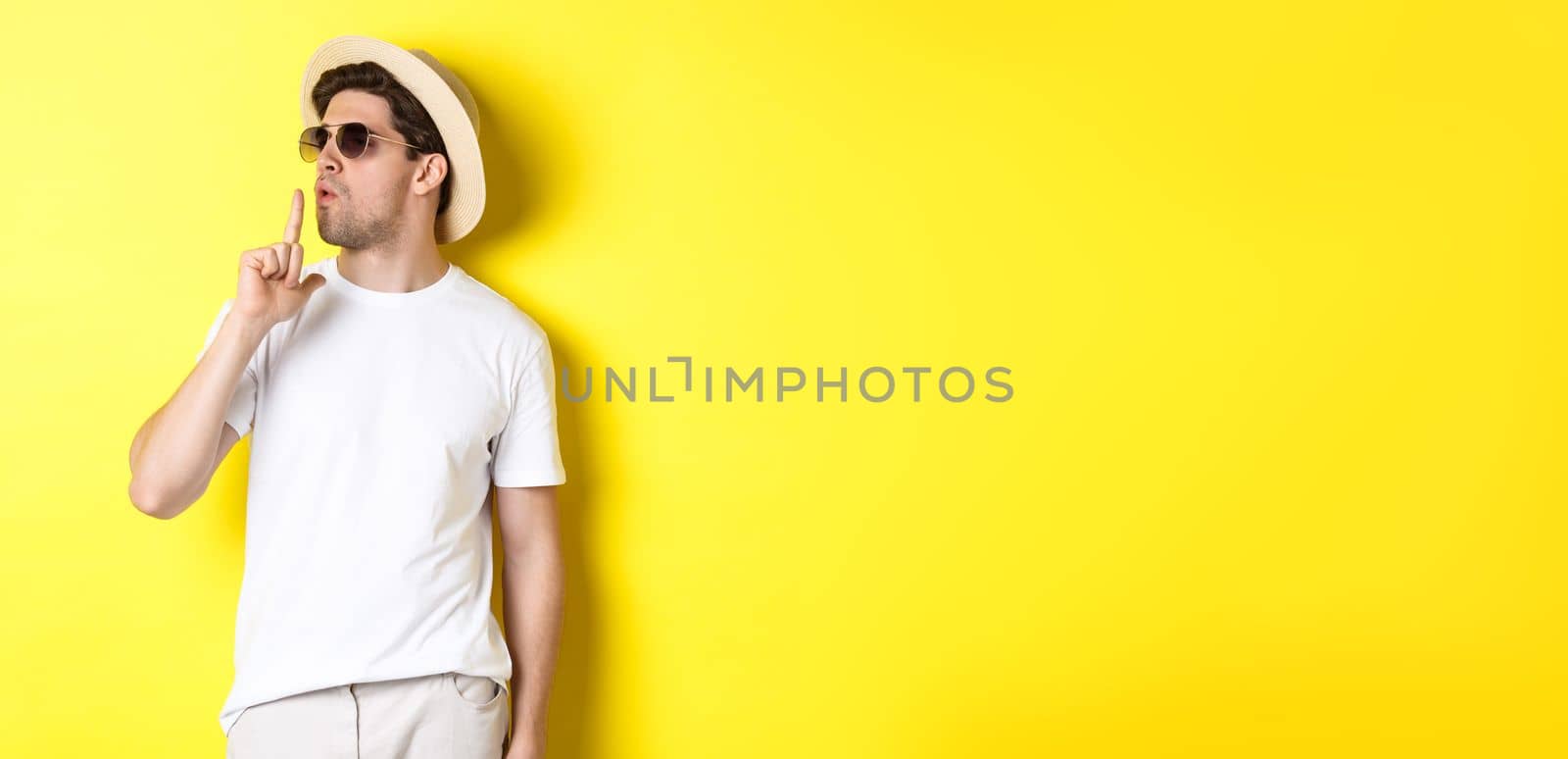 Cool young male tourist blowing at finger gun and looking confident, standing against yellow background. Vacation and lifestyle concept by Benzoix