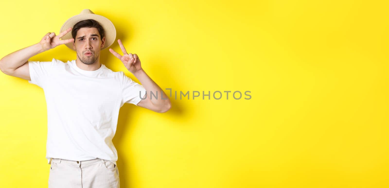 Concept of tourism and vacation. Cool guy taking photo on holidays, posing with peace signs and wearing straw hat, standing against yellow background.