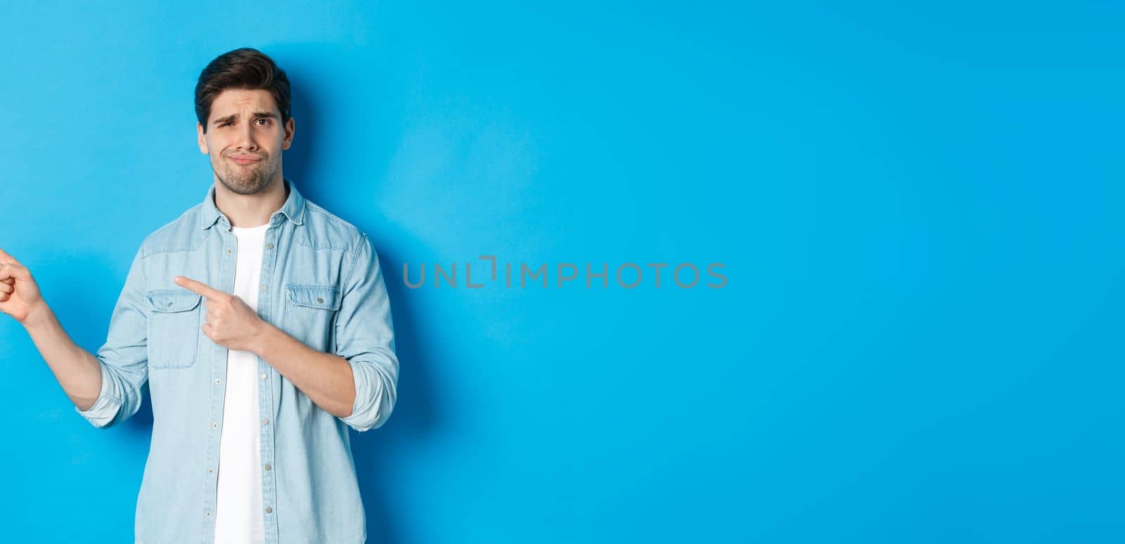 Skeptical adult man pointing fingers left, looking doubtful and unsure, standing over blue background.