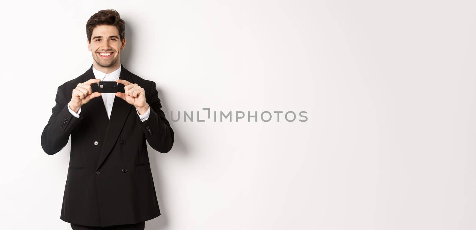 Portrait of confident handsome businessman, smiling and showing credit card, standing in suit against white background.