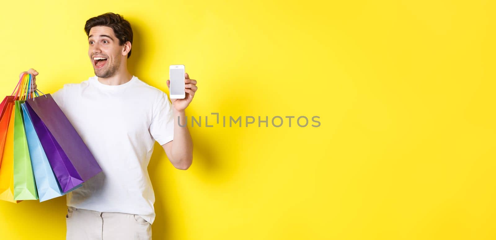 Excited man showing smartphone screen and shopping bags, achieve app goal, demonstrating mobile banking application, yellow background.