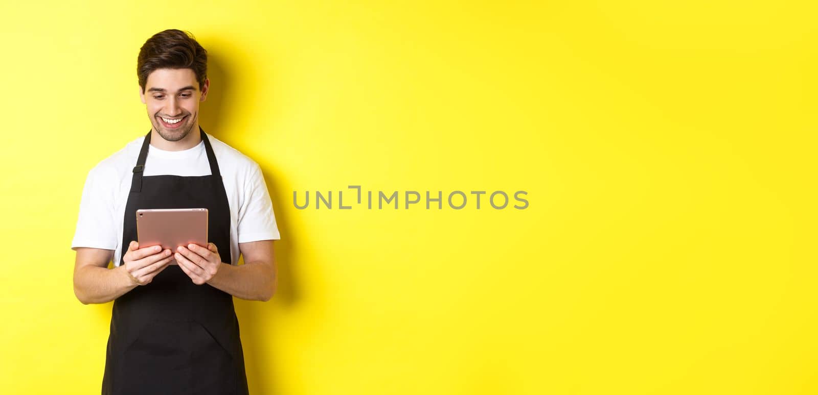 Seller in black apron looking at digital tablet screen, smiling pleased, standing over yellow background by Benzoix