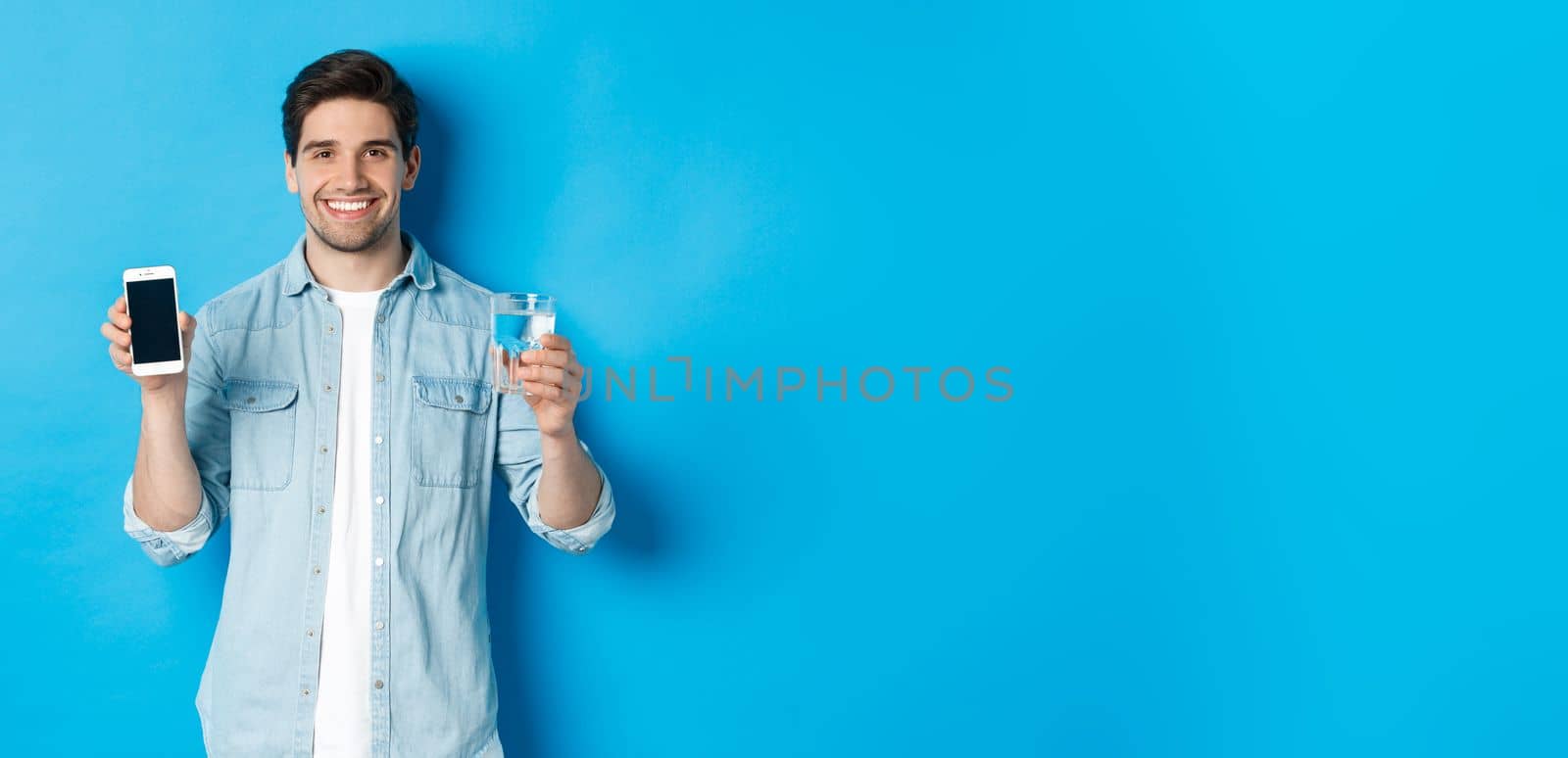 Young man control water balance with smartphone app, showing mobile screen app and smiling, standing over blue background.