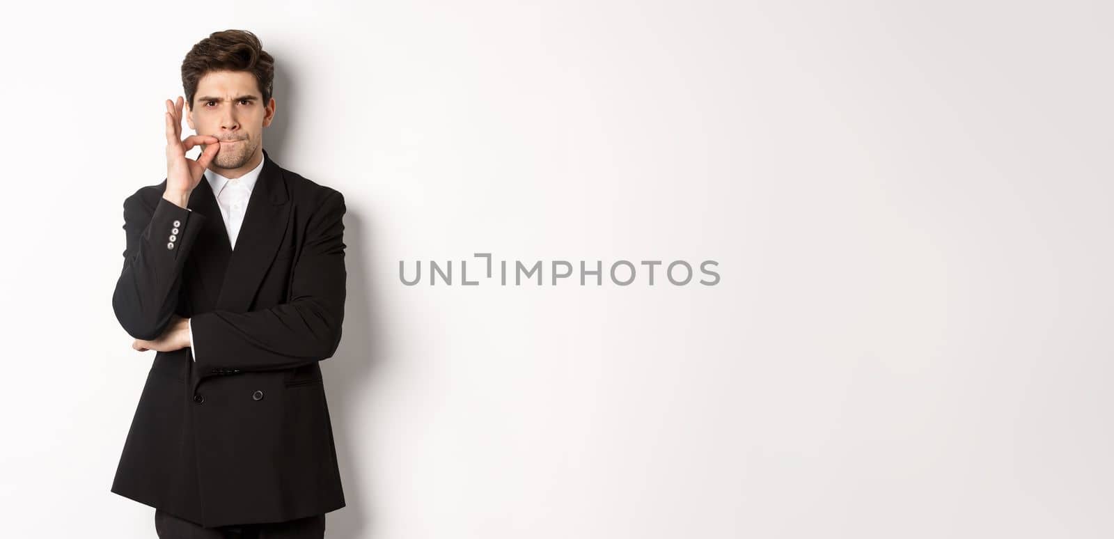 Image of serious businessman in black suit, zipping mouth and frowning, hiding secret, showing taboo sign, standing over white background by Benzoix