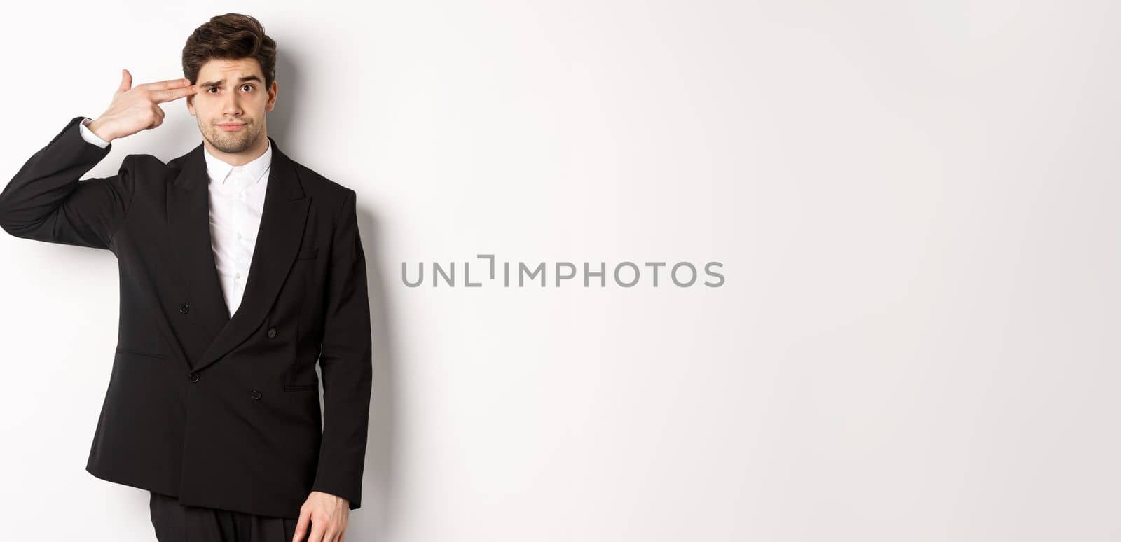 Portrait of distressed and troubled businessman in suit, pointing finger gun at head and looking bothered, shooting himself, standing against white background by Benzoix