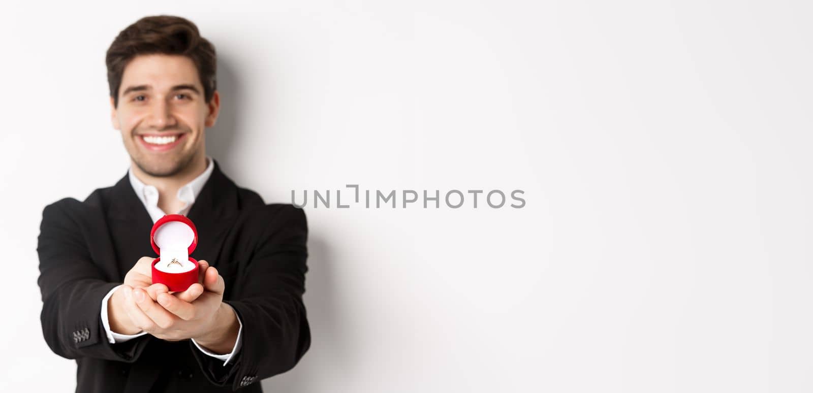 Image of handsome man looking romantic, giving you an engagement ring, making proposal to marry him, standing against white background.