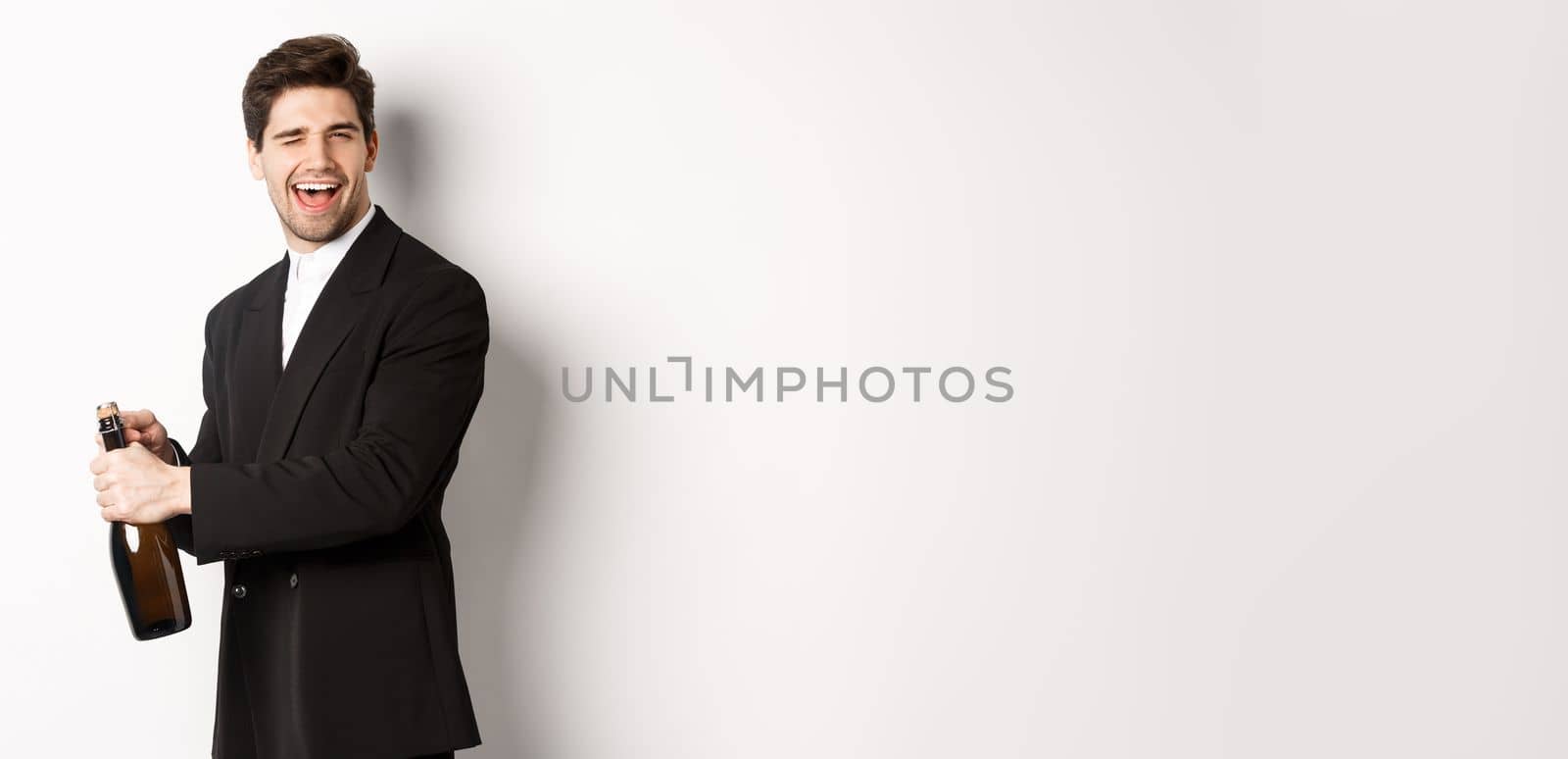 Portrait of attractive man in black suit, winking at camera and opening bottle of champagne, celebrating new year, standing against white background.