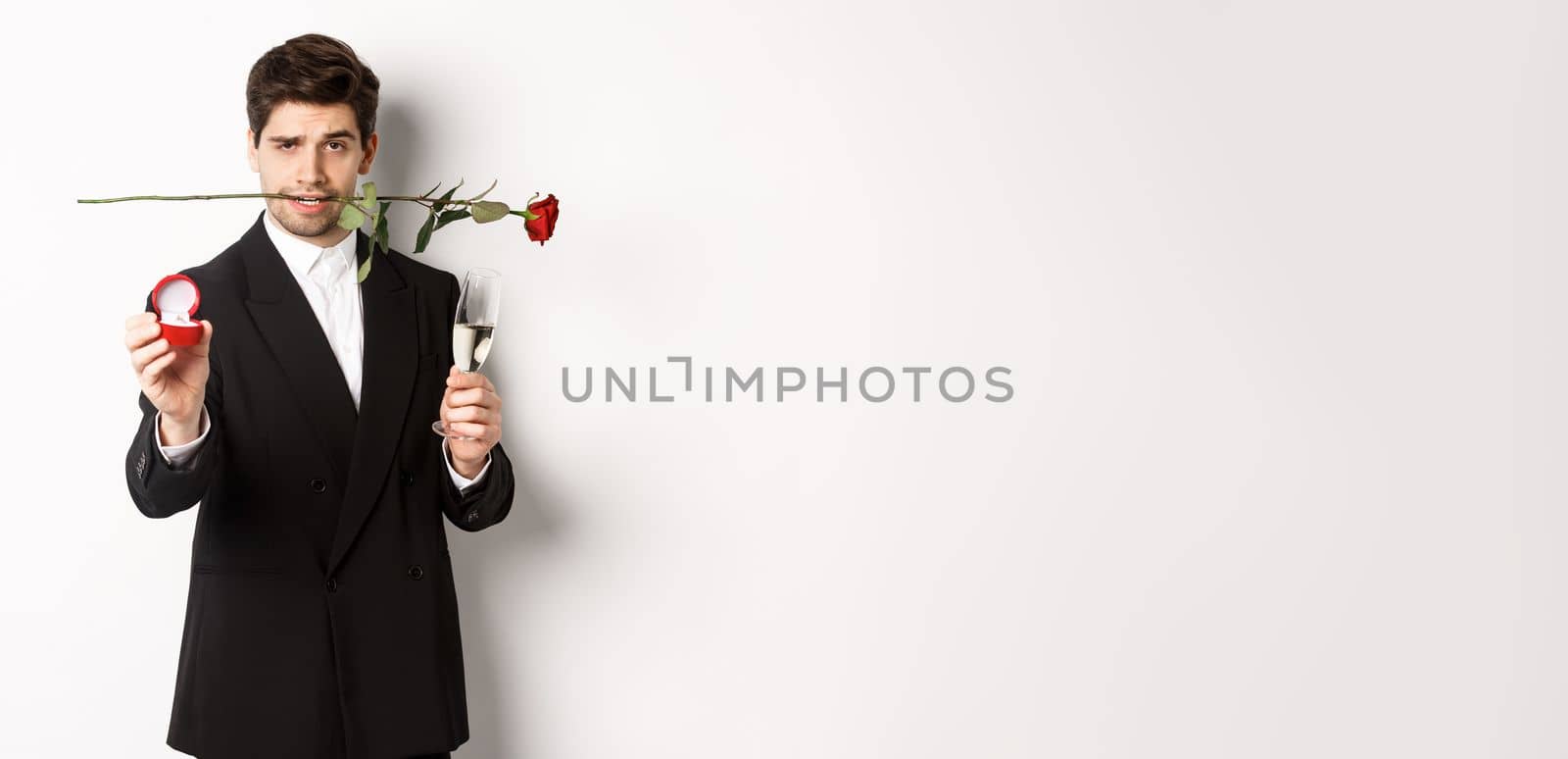 Passionate young man in suit making a proposal, holding rose in teeth and glass of champagne, showing engagement ring, asking to marry him, standing against white background by Benzoix