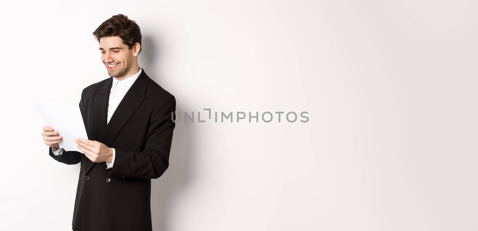 Image of attractive businessman in black suit, reading document and smiling, working on report, standing against white background.