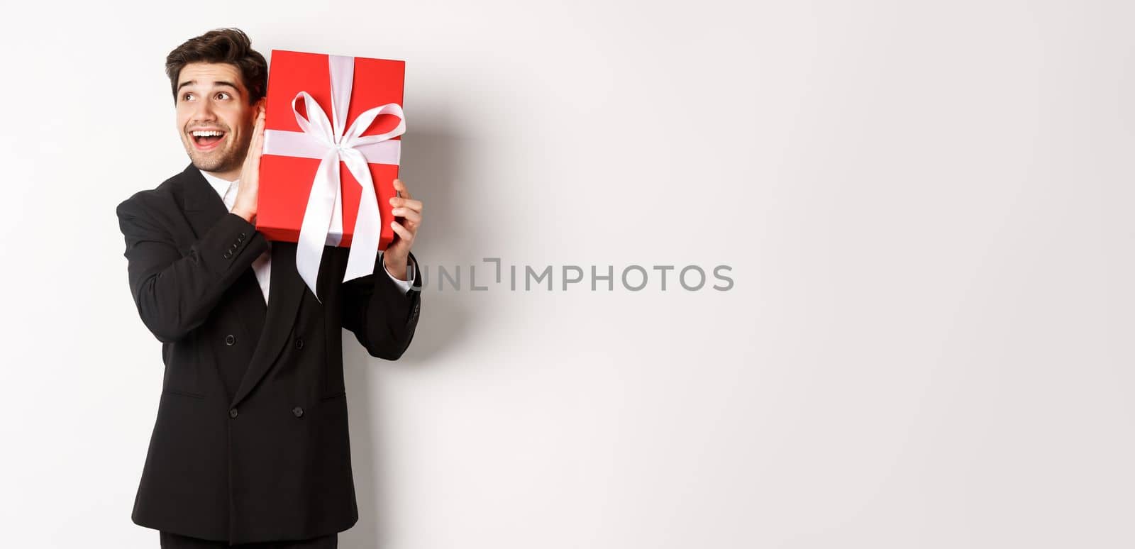 Image of handsome dreamy guy in black suit, shaking box with present to wonder whats inside, standing against white background happy.