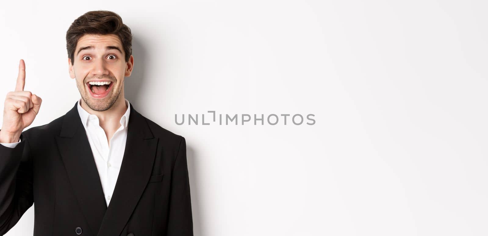 Close-up of handsome businessman in black suit, smiling amazed, showing number one, standing over white background.