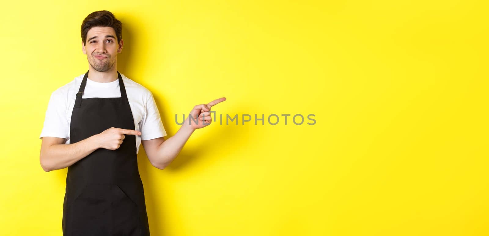 Doubtful barista in black apron pointing fingers right, looking skeptical and unamused, standing over yellow background.