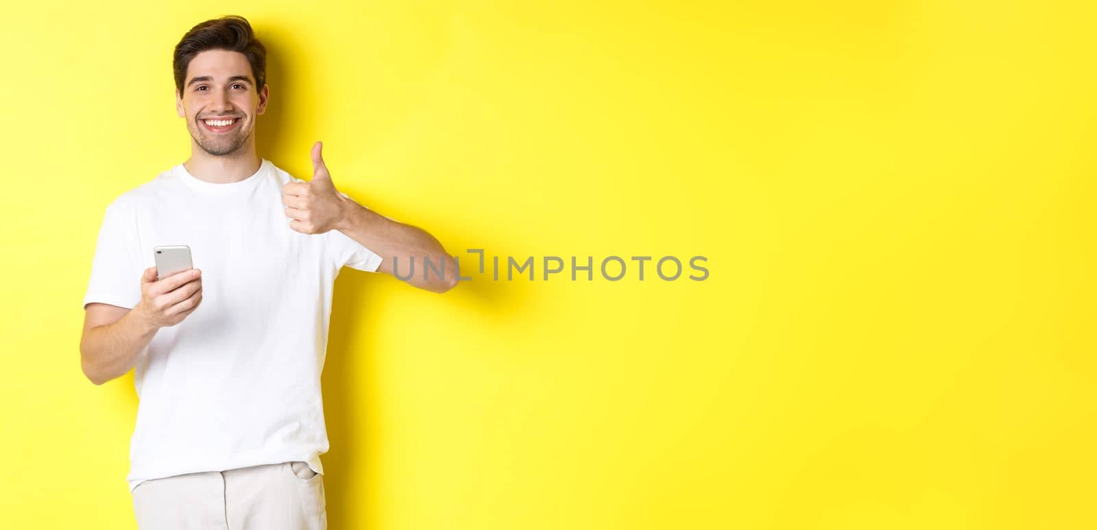 Happy satisfied man holding smartphone, showing thumb up in approval, recommend something online, standing over yellow background.