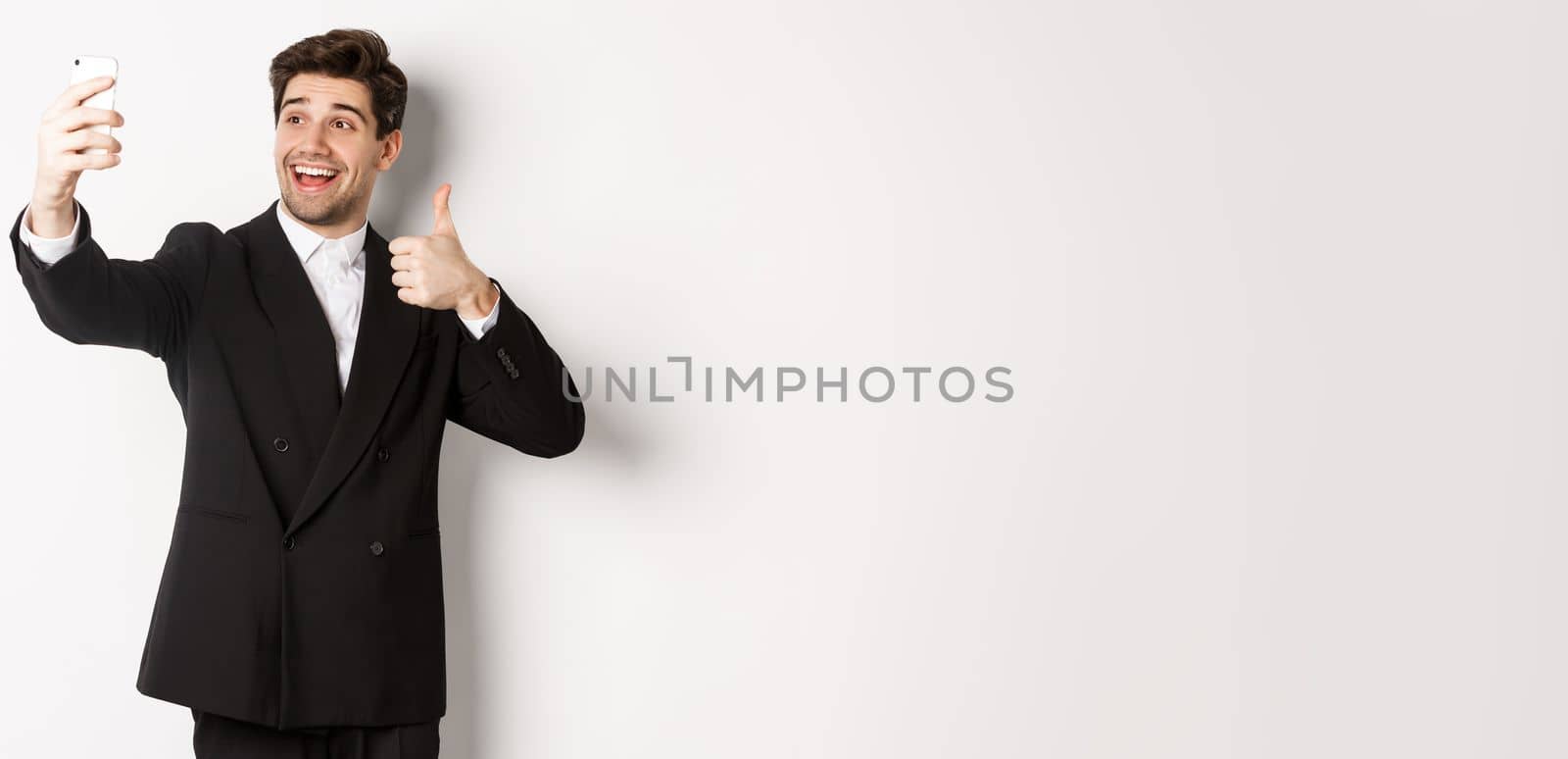 Portrait of good-looking man taking selfie on new year party, wearing suit, taking photo on smartphone and showing thumbs-up, standing against white background by Benzoix