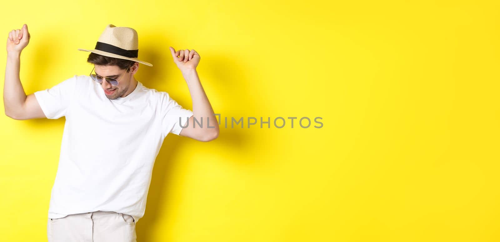 Tourism, travelling and holidays concept. Man tourist enjoying vacation, dancing in straw hat and sunglasses, posing against yellow background.