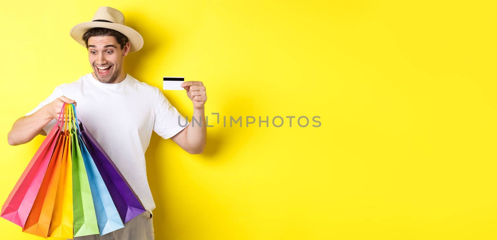 Concept of vacation and finance. Happy man shopper looking at shopping bags satisfied, showing credit card, standing against yellow background by Benzoix
