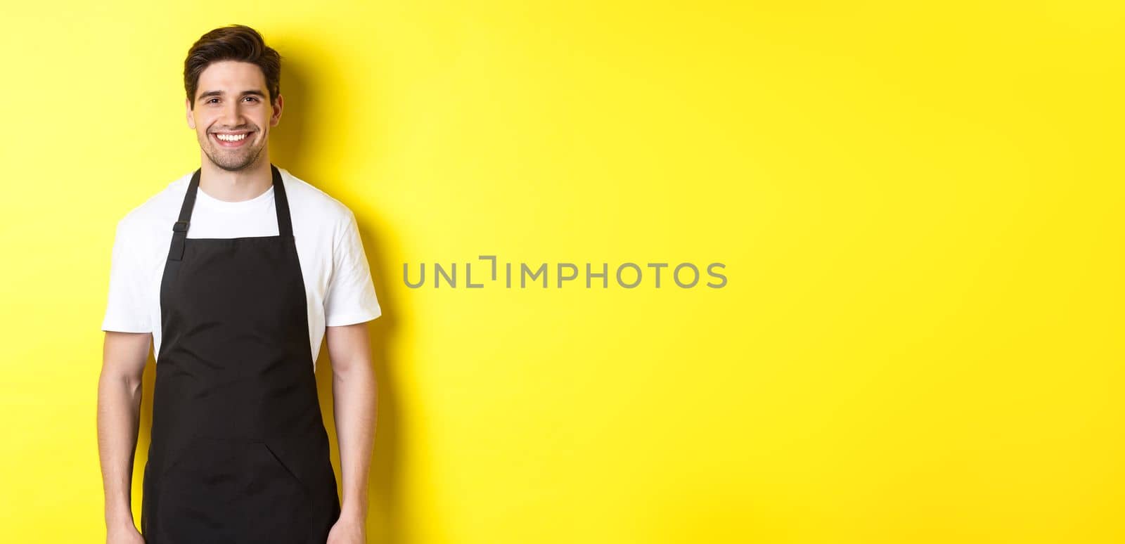 Young man waiter in black apron, smiling, working in store or coffee shop, standing against yellow background.