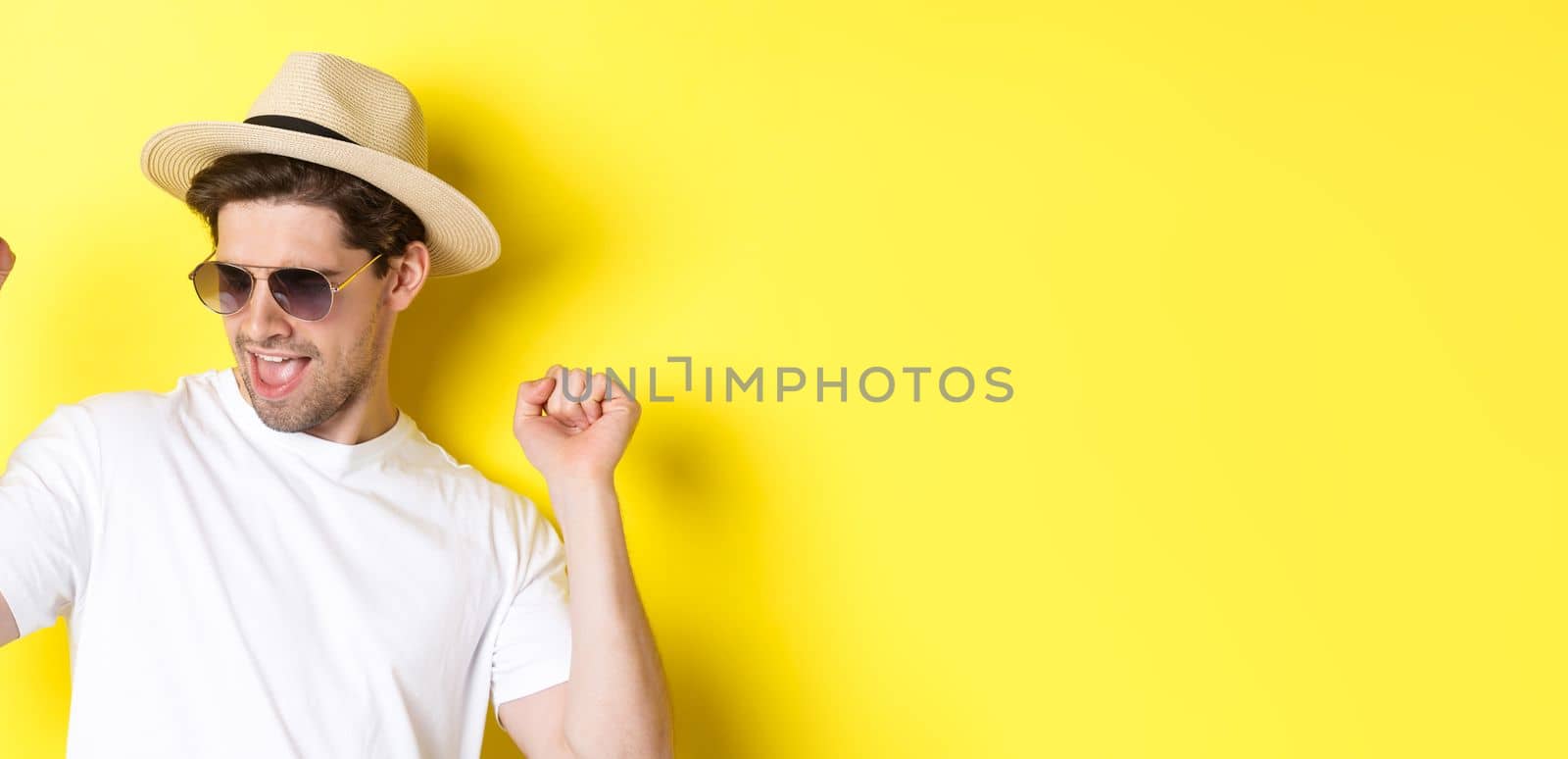 Concept of tourism and vacation. Close-up of man enjoying holidays on trip, dancing and pointing fingers sideways, wearing sunglasses with straw hat, yellow background.