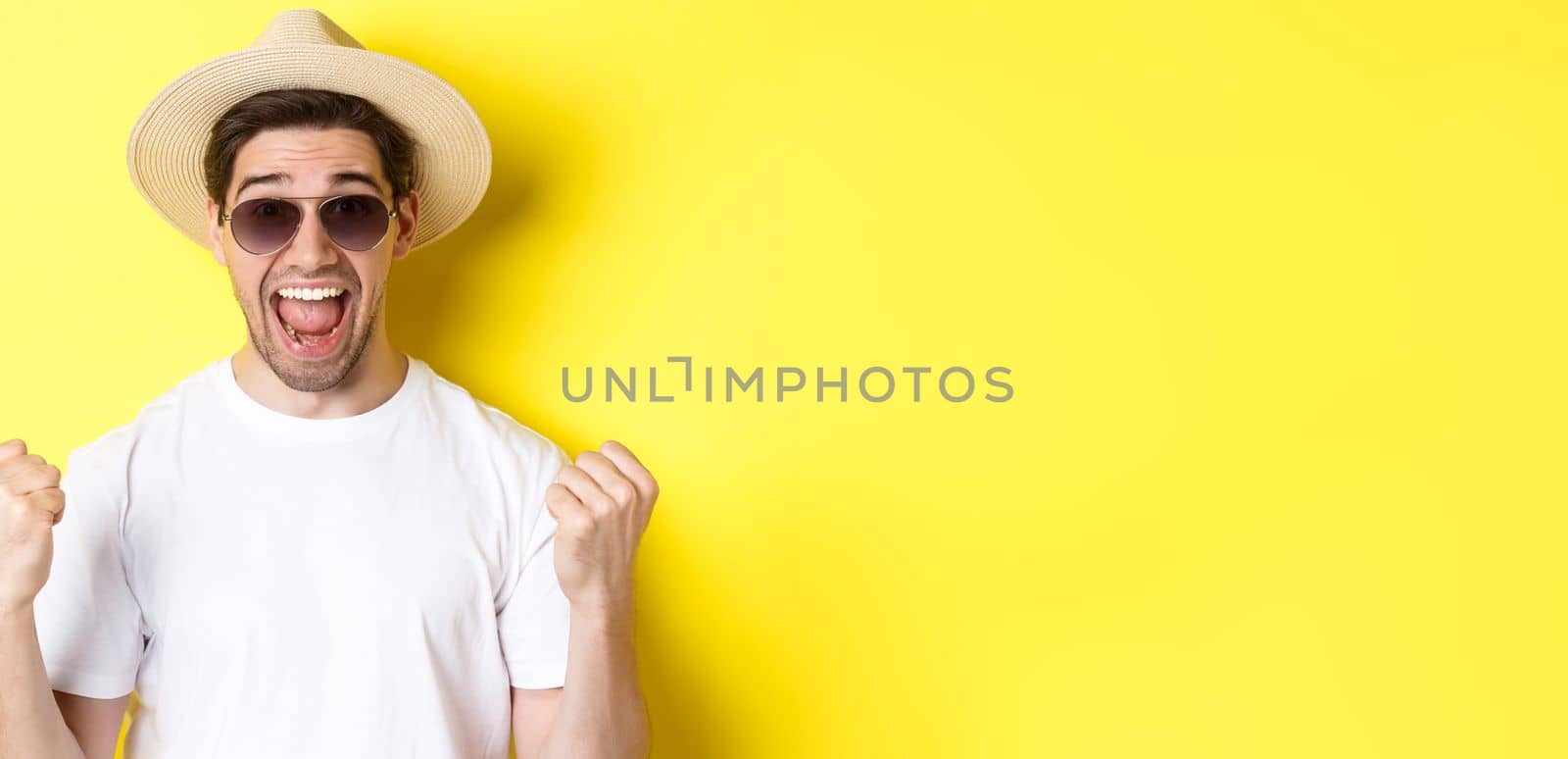 Concept of tourism and holidays. Lucky man tourist winning tickets, making fist pump and saying yes, wearing vacation clothes, standing over yellow background.