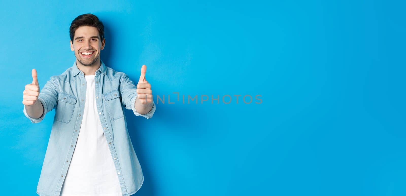 Smiling confident man showing thumbs up, guarantee quality, approving something good, standing against blue background.