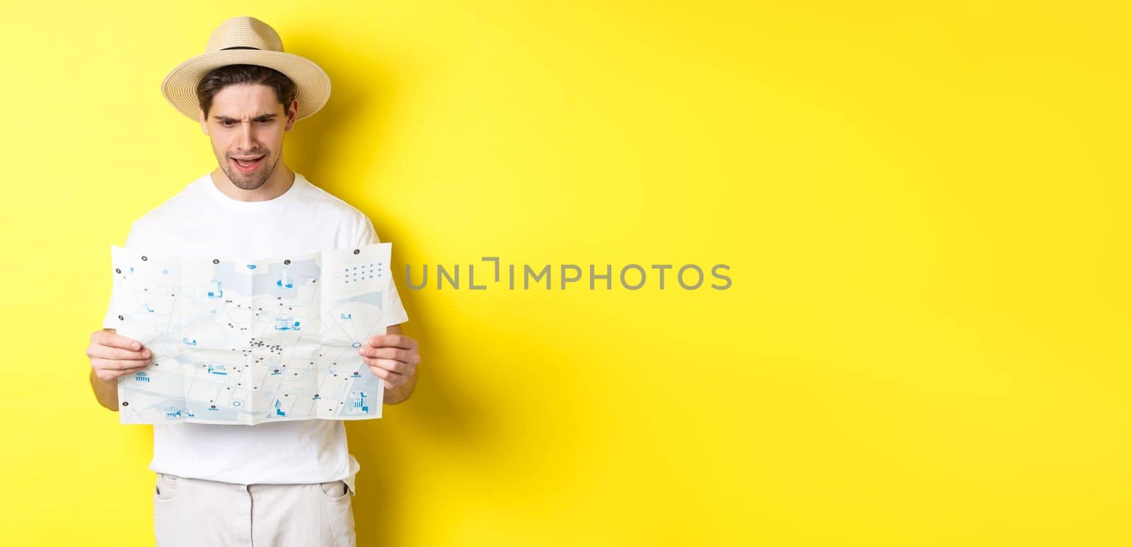 Travelling, vacation and tourism concept. Man looking confused at map during trip, cant understand, standing over yellow background.