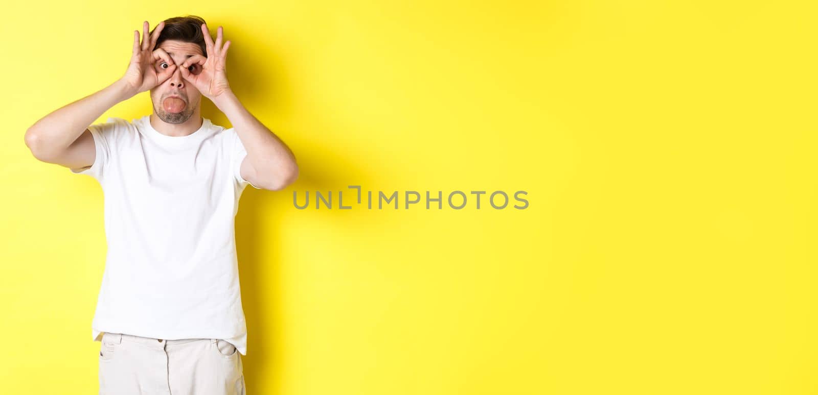 Young man making funny faces and showing tongue, fool around, standing in white t-shirt against yellow background by Benzoix