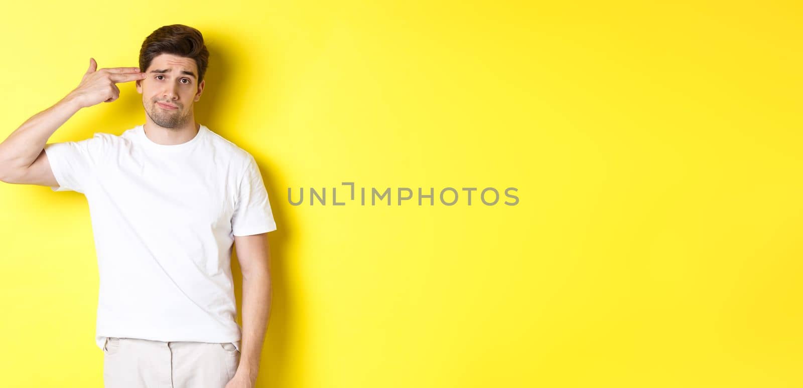 Bothered man shooting himself with finger gun, looking displeased and tired, standing against yellow background.
