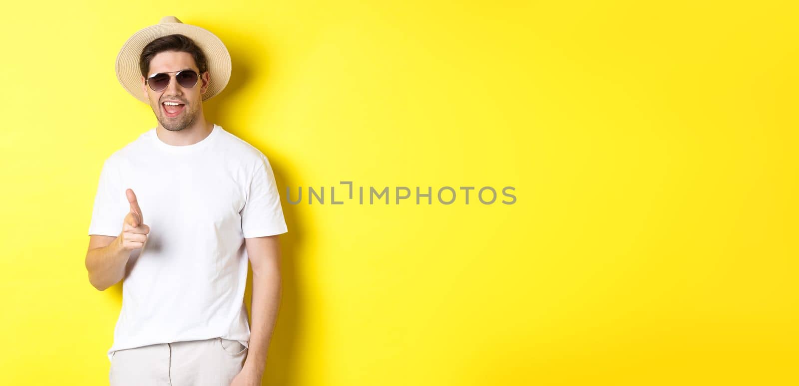 Confident and cheeky guy on vacation flirting with you, pointing finger at camera and winking, wearing summer hat with sunglasses, yellow background.