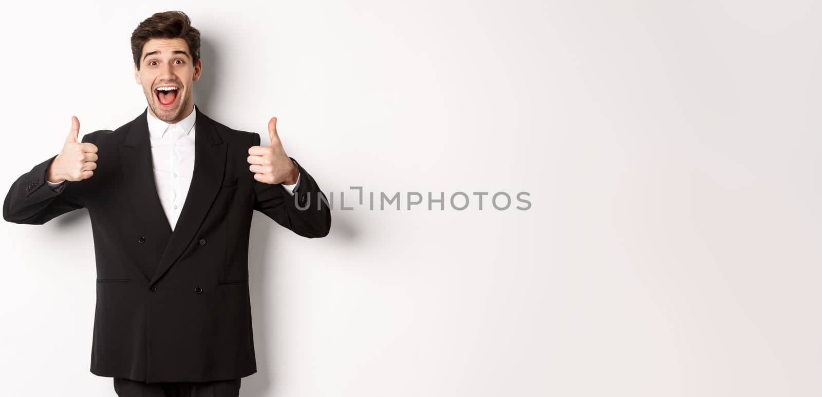 Concept of new year party, celebration and lifestyle. Portrait of amazed and pleased handsome man in black suit, showing thumbs-up, like product, approve something good, white background.