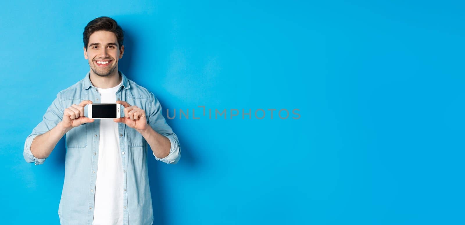 Handsome smiling man showing smartphone screen, standing against blue background for copy space.