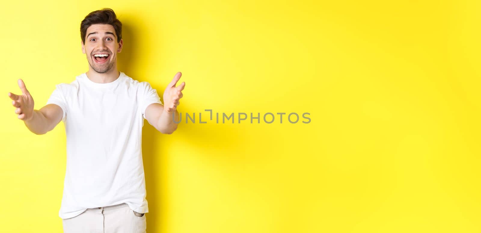 Excited handsome guy stretching hands forward, reaching for hug, receiving gift, standing over yellow background by Benzoix