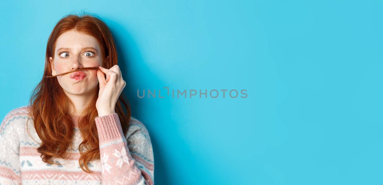 Close-up of silly and funny redhead girl making moustache with hair strand and puckered lips, grimacing against blue background.