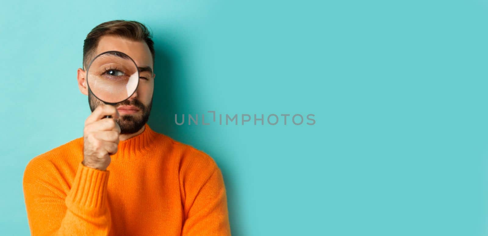 Funny man looking through magnifying glass, searching or investigating something, standing in orange sweater against turquoise background.