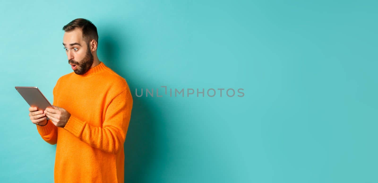 Image of male model in orange sweater staring at digital tablet screen, looking surprised, standing over light blue background by Benzoix
