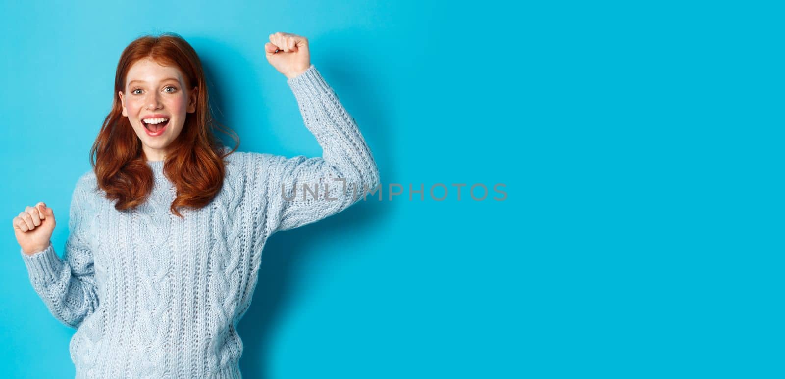 Cheerful redhead gil winning, celebrating victory, smiling and jumping from happiness, posing against blue background.