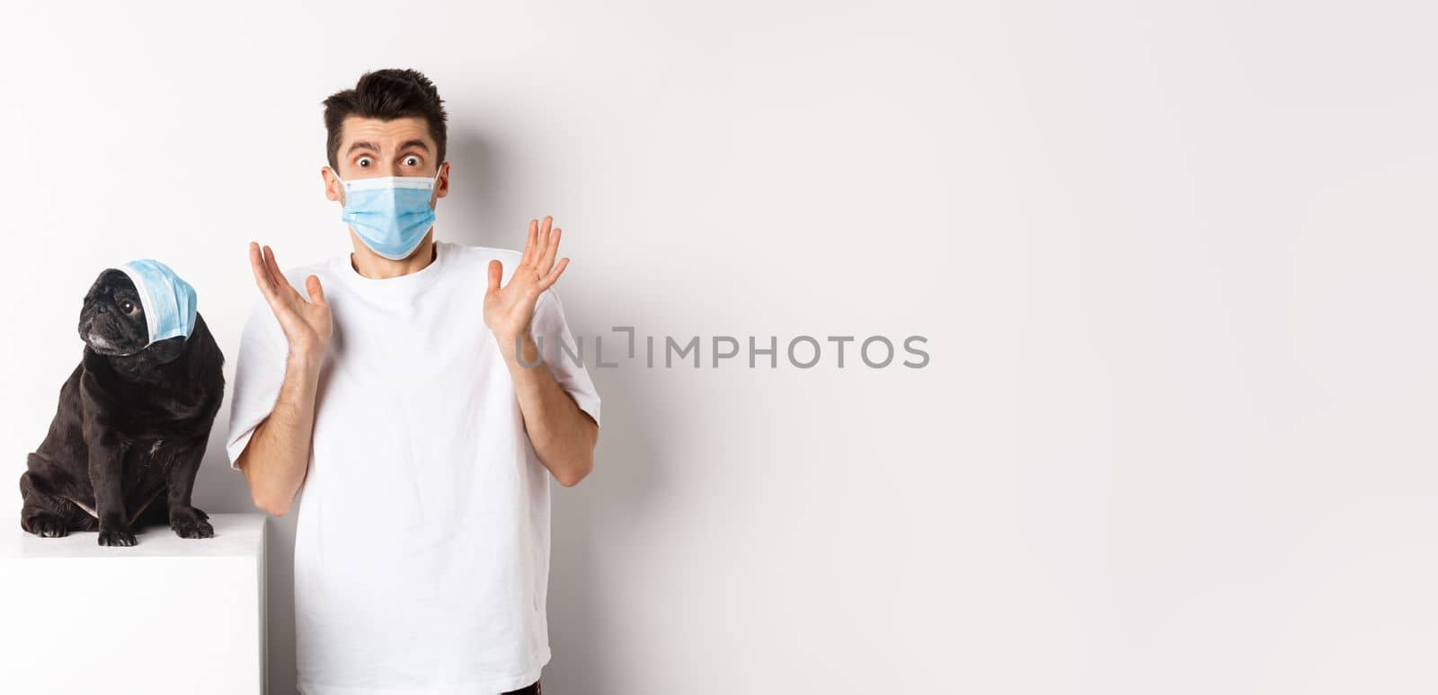 Covid-19, animals and quarantine concept. Shocked dog owner and pug wearing medical masks, staring at camera amazed, standing over white background.