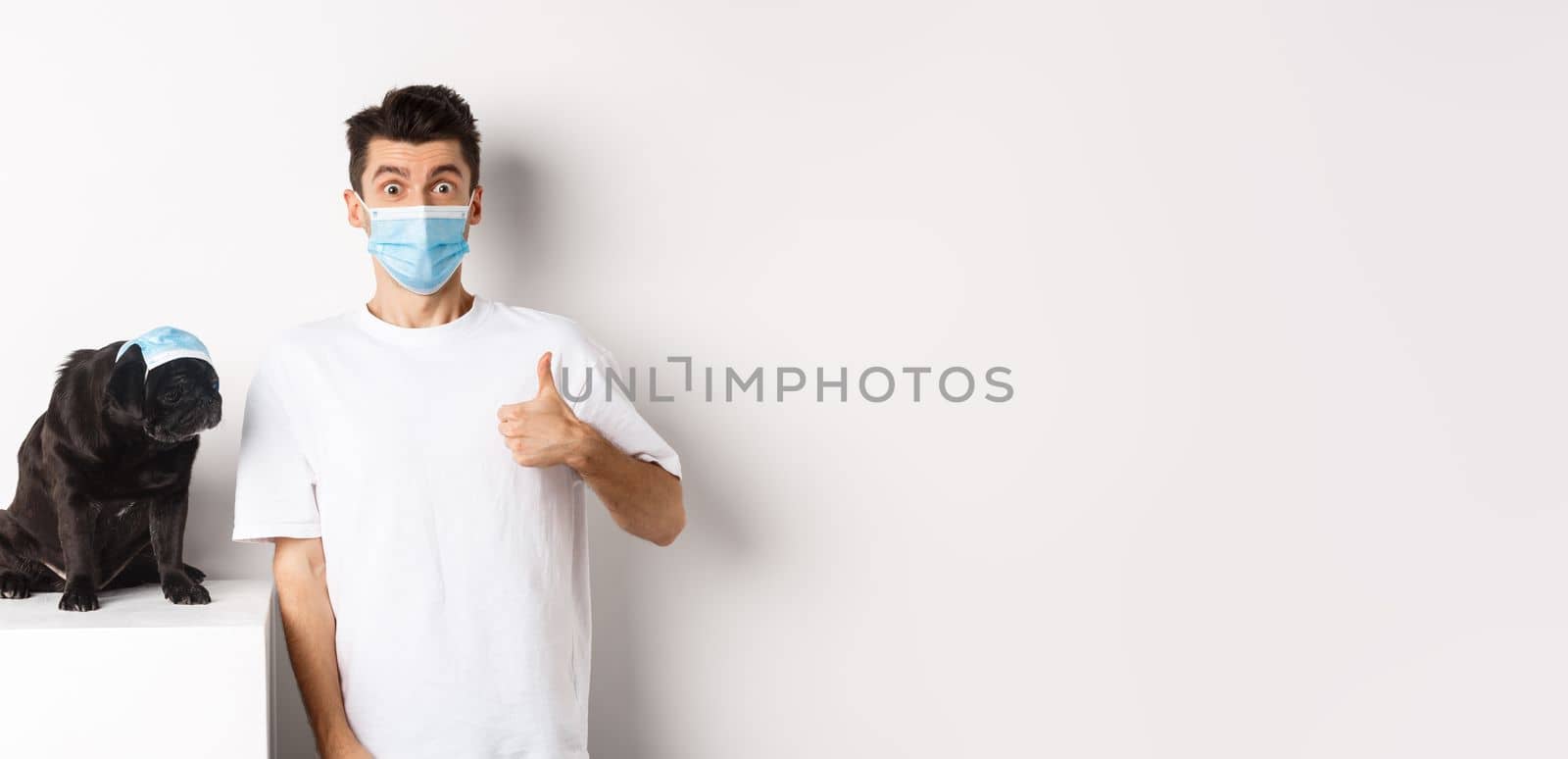 Covid-19, animals and quarantine concept. Handsome young man and small dog wearing face masks, owner showing thumb up in approval, praise super cool product, white background by Benzoix