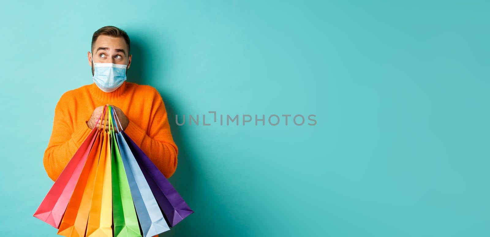 Covid-19, pandemic and lifestyle concept. Man thinking, wearing face mask, holding shopping bags, imaging something, standing over turquoise background by Benzoix