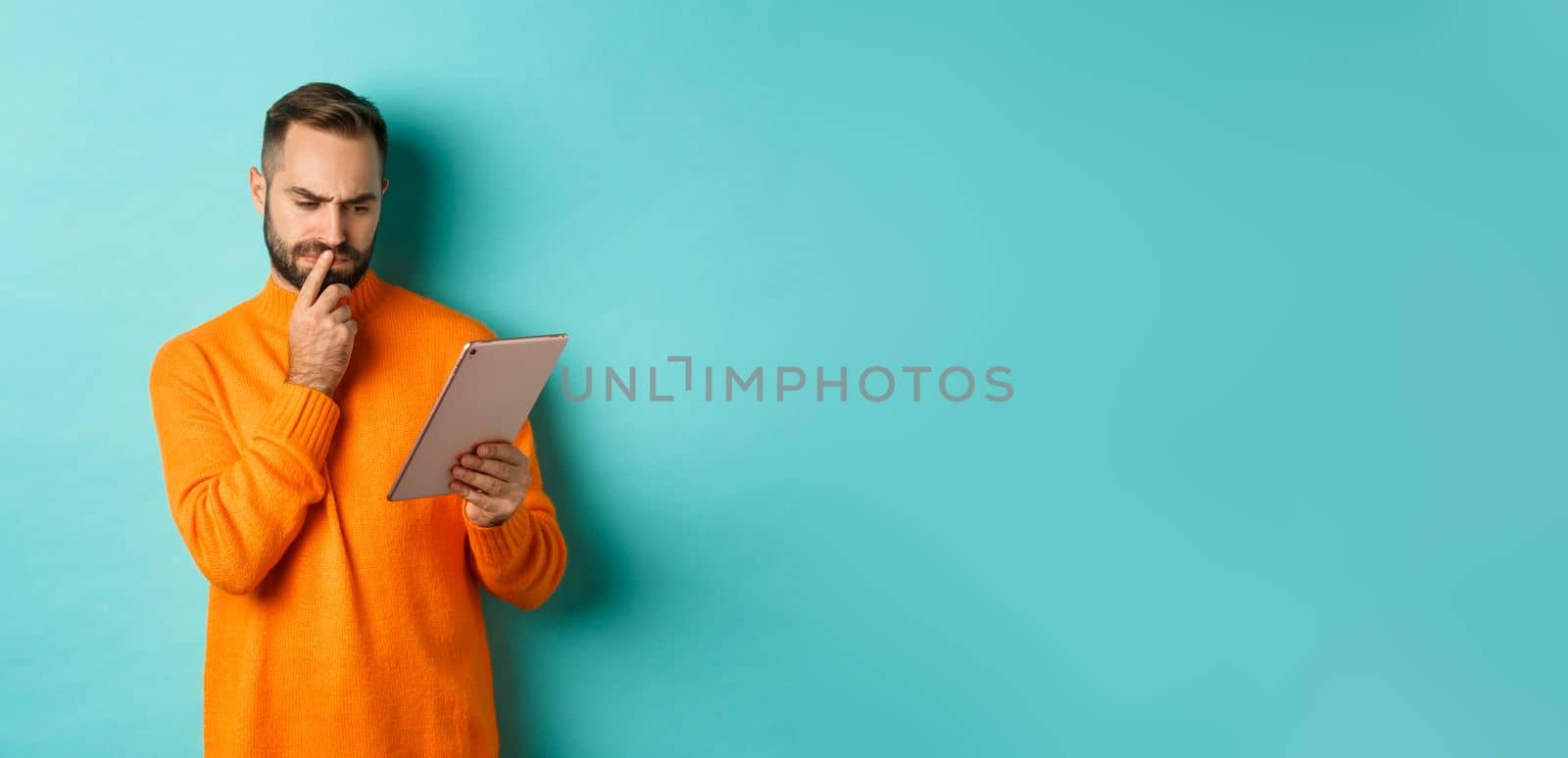 Serious man pondering and looking at digital tablet screen, reading social media, standing over turquoise background. Copy space