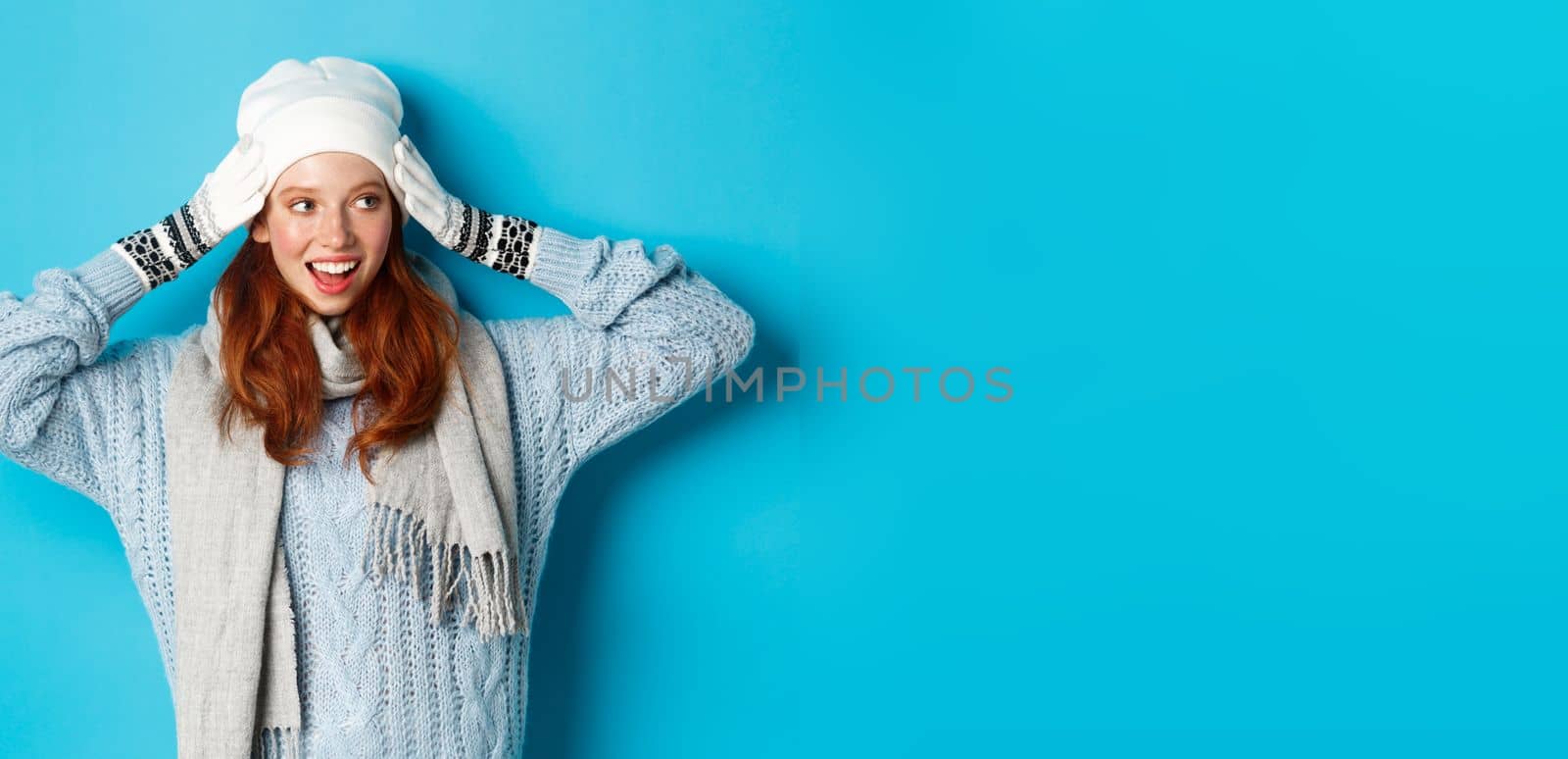 Winter and holidays concept. Happy redhead girl in beanie hat, scarf and gloves looking right and smiling, standing against blue background.