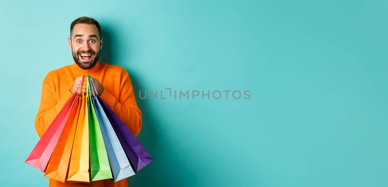 Excited adult man holding shopping bags and smiling, going to mall, standing over turquoise background.