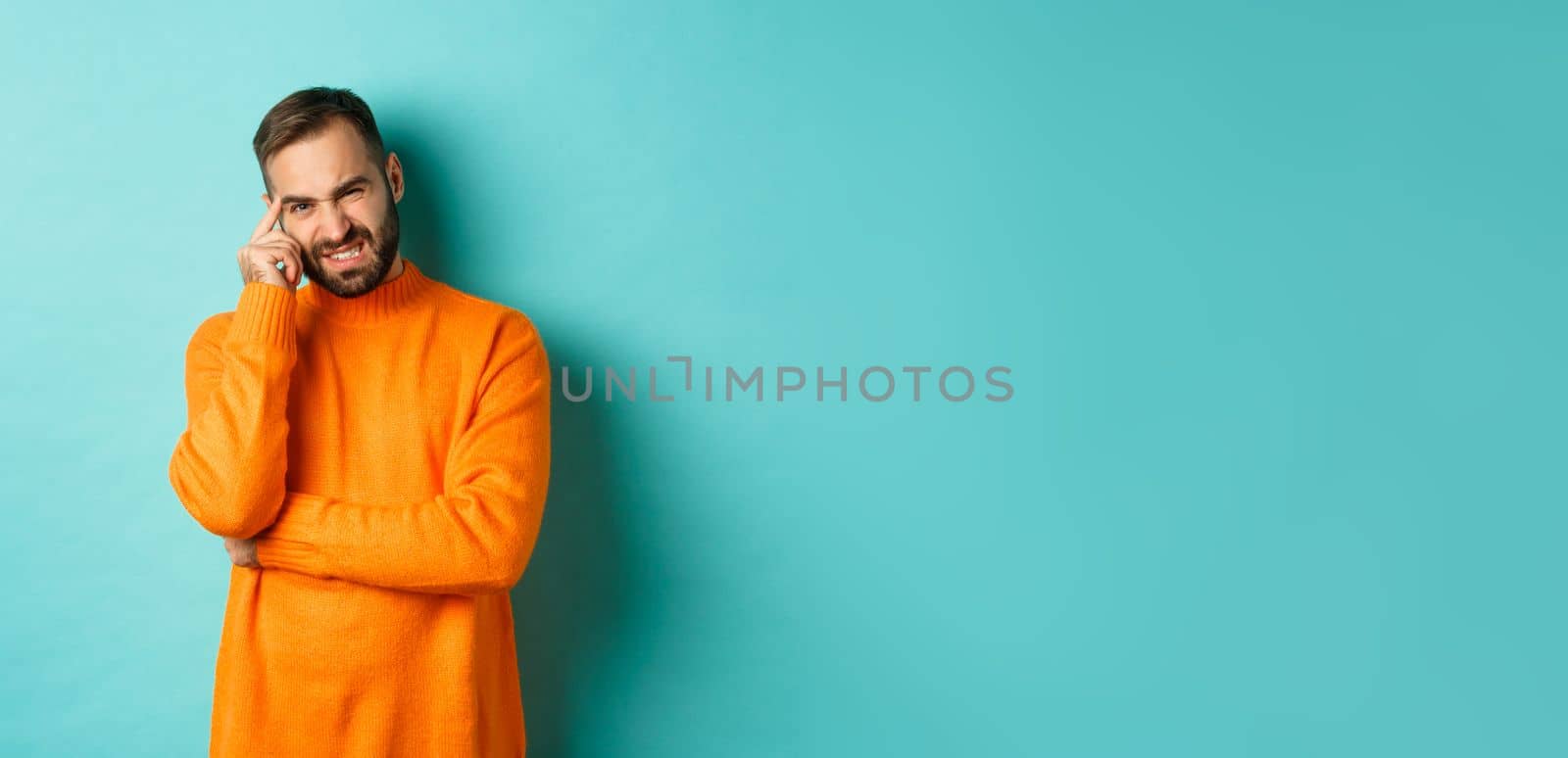 Man with beard, grimacing and cringe from something embarrassing, looking with awkward face, standing over light blue background.