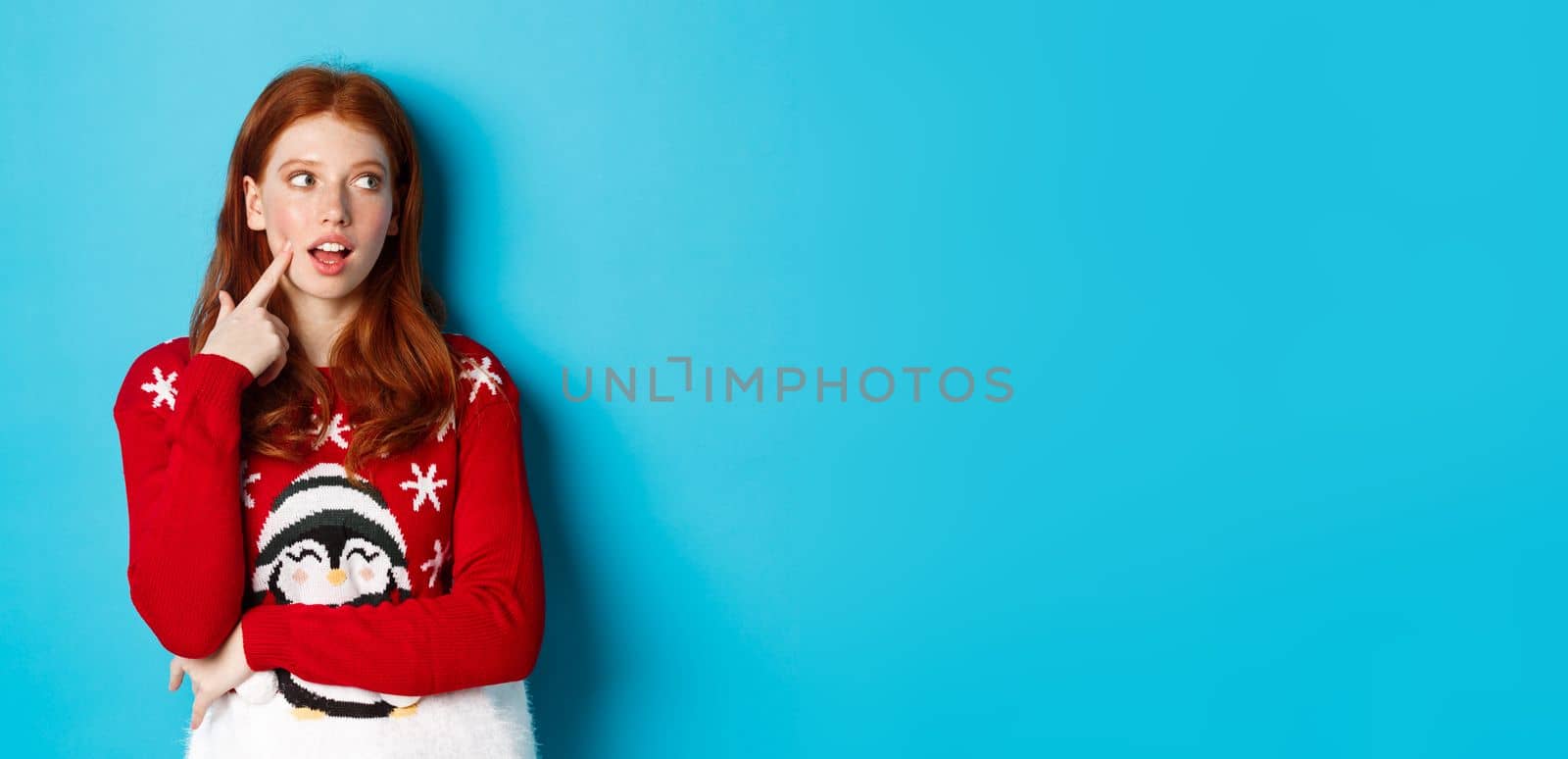 Winter holidays and Christmas Eve concept. Pretty redhead girl in xmas sweater, touching cheek thoughtful and smiling, making choice, looking at upper right corner and thinking, blue background.