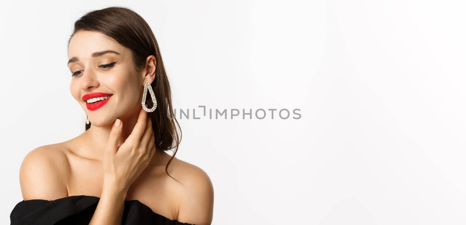 Fashion and beauty concept. Close-up of tender woman in black dress and earrings, gently touching face and smiling, looking down coquettish, standing over white background.