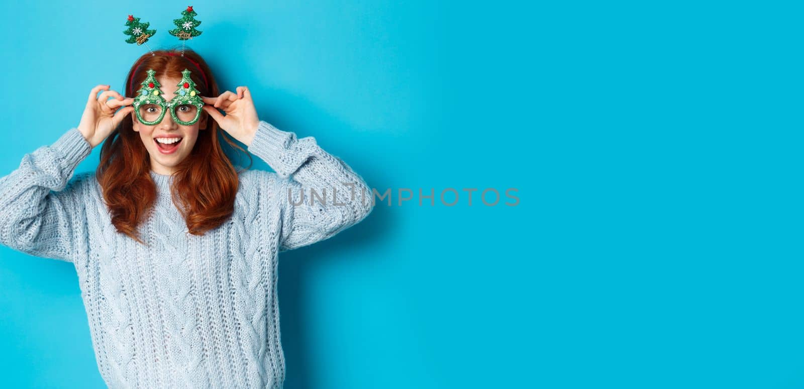 Winter holidays and Christmas sales concept. Beautiful redhead female model celebrating New Year, wearing funny party headband and glasses, smiling silly, blue background by Benzoix