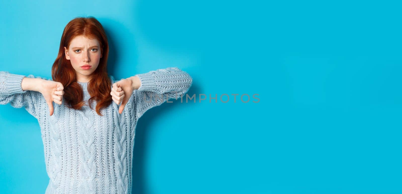 Disappointed redhead girl in sweater showing thumbs-down, judging bad product, disagree and dislike promo, standing over blue background.